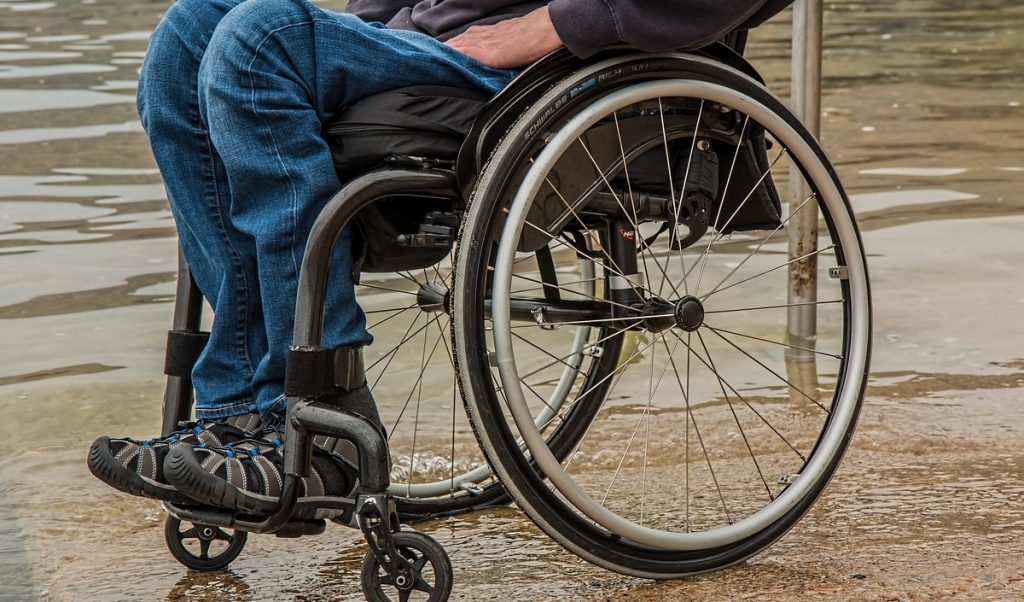 Descripción gráfica: sobre un fondo café, persona en silla de ruedas, la cual se observa desde la cintura hacia abajo, enfocando en gran parte la silla de ruedas en primer plano.