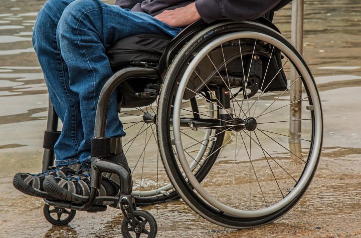 Descripción gráfica: sobre un fondo café, persona en silla de ruedas, la cual se observa desde la cintura hacia abajo, enfocando en gran parte la silla de ruedas en primer plano.